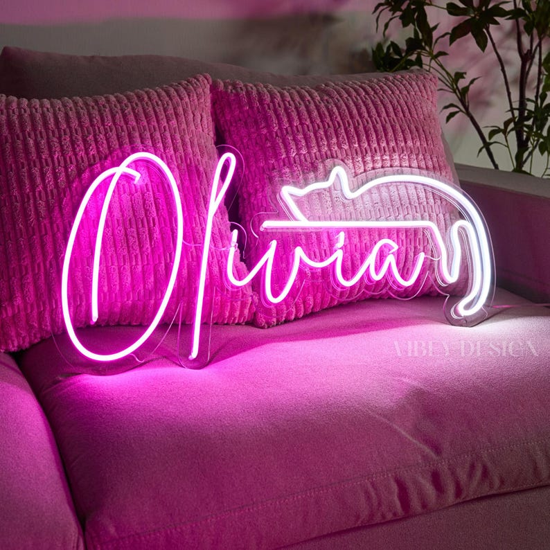 K&ouml;nnte beinhalten: Ein leuchtend pinkes Neonschild, das den Namen Olivia in Schreibschrift zeigt, integriert mit der stilisierten Umrissform einer Katze. Die Neonr&ouml;hre ist auf einer transparenten Acrylplatte montiert und wirft einen lebendigen Schein auf die texturierten pinken Kissen.