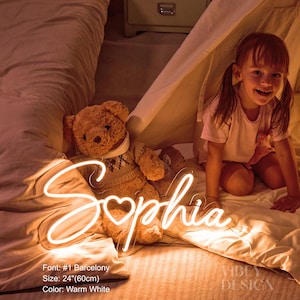 K&ouml;nnte beinhalten: Ein warmwei&szlig;es Neonschild mit dem Namen Sophia in Schreibschrift, bei dem ein Herz den Buchstaben o ersetzt. Das Schild ist 60 cm breit, im Barcelony-Schriftstil gehalten und strahlt neben einem Teddyb&auml;ren ein sanftes Licht aus.