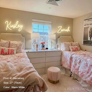 K&ouml;nnte beinhalten: Zwei individuelle, warmwei&szlig;e Neonreklamen an der Wand &uuml;ber zwei Einzelbetten, die die Namen Kinsley und Zariah in Schreibschrift zeigen. Das Schlafzimmer ist mit Blumenbettw&auml;sche, zwei Nachttischlampen und einem kleinen rosa Polsterhocker ausgestattet.