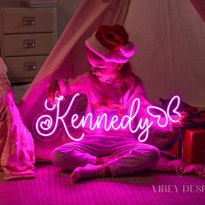 K&ouml;nnte beinhalten: Ein individuelles Neonschild mit dem Namen Kennedy in leuchtend pinker Schreibschrift, verziert mit einem kleinen Herz auf der linken Seite und einer Schmetterlingskontur auf der rechten Seite. Das Schild ist auf Acryl montiert und leuchtet pink.