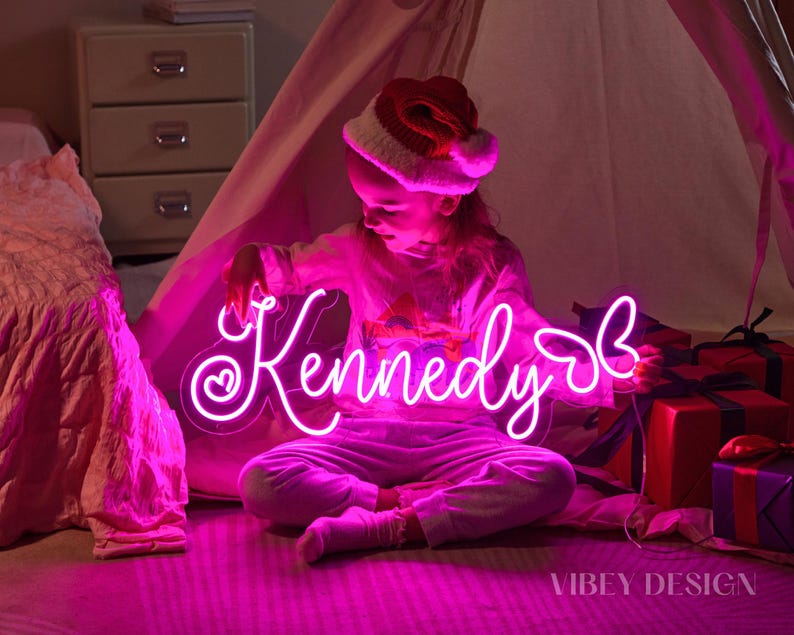 K&ouml;nnte beinhalten: Ein individuelles Neonschild mit dem Namen Kennedy in leuchtend pinker Schreibschrift, verziert mit einem kleinen Herz auf der linken Seite und einer Schmetterlingskontur auf der rechten Seite. Das Schild ist auf Acryl montiert und leuchtet pink.