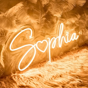 Może przedstawiać: Biały neonowy znak z imieniem "Sophia" w ozdobnym, pochylonym stylu pisma z małym sercem w środku imienia. Znak znajduje się na puszystej, jasnobrązowej powierzchni.
