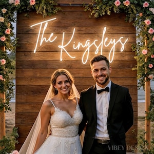 Puede incluir: Escena de boda con una pareja sonriendo frente a un fondo de madera. Un letrero de neón dice "The Kingsleys". La novia lleva un vestido de novia de encaje blanco y velo. El novio viste un traje negro con pajarita. Arreglos florales enmarcan el fondo.