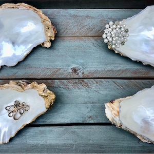 May include: Four white oyster shells with gold trim, displayed on a wooden surface. Each shell is filled with a different decorative item, including a pearl brooch, a silver octopus charm, and two other decorative items.