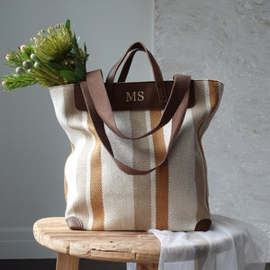 May include: A striped canvas tote bag with brown leather handles and the initials "MS" embossed on the front. The bag is sitting on a wooden stool with a white cloth draped over it.