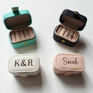 May include: Four small ring boxes in different colours. The boxes are made of faux leather and have a hinged lid. The boxes are personalised with the names "K&R" and "Sarah". One box has a ring inside.