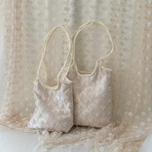 Pode incluir: Duas sacolas creme com bordados florais. As sacolas têm alças longas e finas e são colocadas em um fundo de tecido correspondente com um padrão floral semelhante. As sacolas são de tamanhos diferentes.