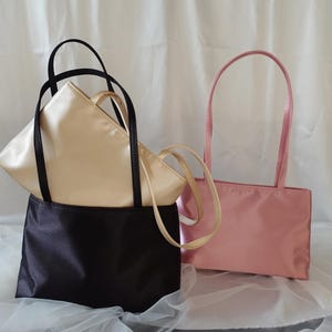 May include: A black handbag with two thin black handles, a cream-colored handbag with two thin cream handles, and a pink handbag with two thin pink handles. Each bag features a rectangular base and a smooth, satin-like finish with a sheen.