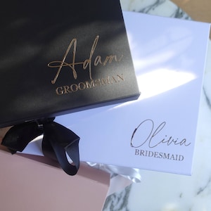 May include: Two gift boxes with black ribbon bows. The black box has "Adam Groomman" written in gold lettering. The white box has "Olivia Bridesmaid" written in gold lettering.