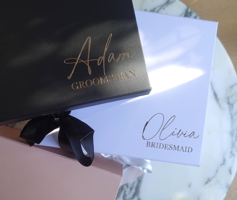 May include: Two gift boxes with black ribbon bows. The black box has "Adam Groomman" written in gold lettering. The white box has "Olivia Bridesmaid" written in gold lettering.
