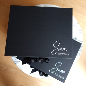 May include: Two black gift boxes with satin ribbon bows. The top box has the name "Sam" written in white script with the words "Best Man" underneath. The bottom box has the name "Saz" written in white script with the words "Maid of Honour" underneath.