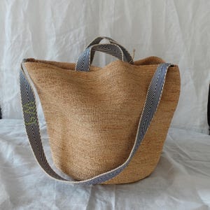 May include: A woven tote bag with a natural tan color. The bag has a blue and white striped shoulder strap and handles. The strap has green embroidered text. The bag is made of a woven material.