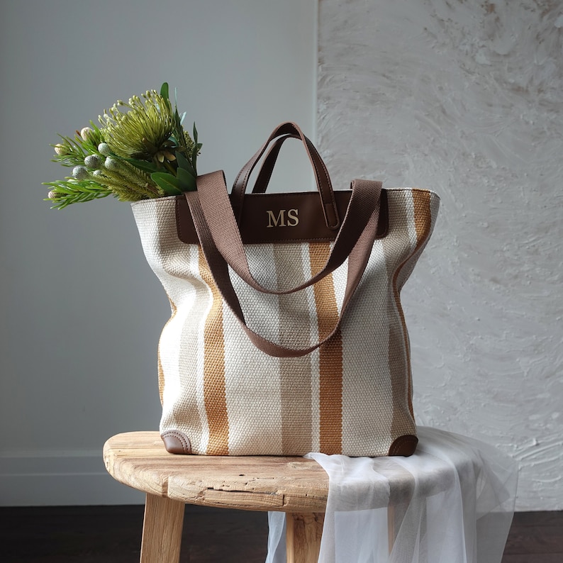 May include: A striped canvas tote bag with brown leather handles and the initials "MS" embossed on the front. The bag is sitting on a wooden stool with a white cloth draped over it.