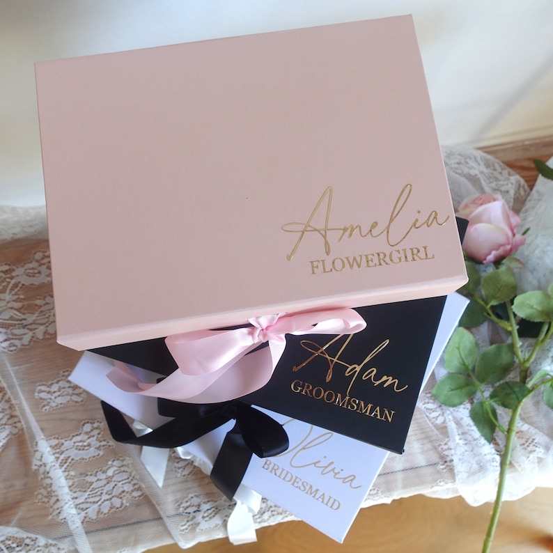 May include: Three gift boxes with pink, black and white lids. The boxes are tied with pink and black ribbons. The lids are personalised with the names "Amelia Flowergirl", "Adam Groomsman", and "Olivia Bridesmaid" in gold lettering.
