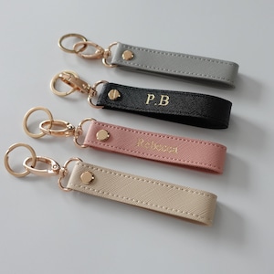 May include: Four rectangular keychains with rose gold hardware, including a lobster clasp and split ring. The straps are made of textured material in gray, black, pink, and beige. The black strap features the gold embossed text P.B, and the pink strap features the gold embossed text Rebecca.