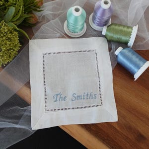 May include: A white linen cocktail napkin with a decorative stitched border and the embroidered text "The Smiths" in light blue. Spools of pastel-coloured thread in blue, green, and purple are arranged around the napkin.