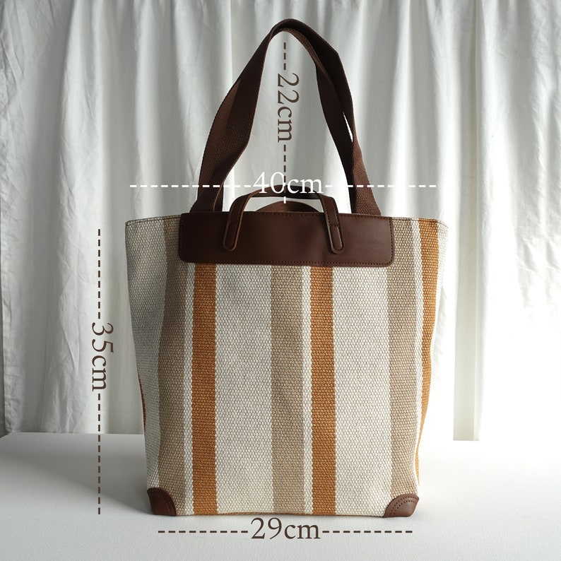 May include: A striped canvas tote bag with brown leather handles and trim. The bag measures 13.75 inches wide, 15.75 inches tall, and 11.4 inches deep.