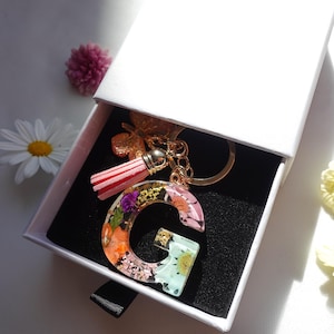 May include: A clear resin letter "G" keyring filled with dried flowers in orange, pink, and purple. The keyring has a gold-coloured chain and tassel, presented in a white gift box.