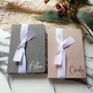 May include: Two personalised grey and pink linen journals with white ribbon bows and the names "Aston" and "Cindy" printed on the covers.