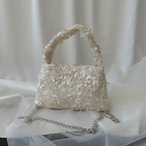 May include: An off-white sequined handbag with a short handle and a silver chain strap. The bag is set against a backdrop of white, draped fabric.