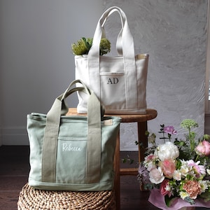 May include: Two canvas tote bags, one off-white and one sage green, with matching handles and front pockets. The off-white bag has the initials "AD" and the green bag has the name "Rebecca" embroidered on the front. Both bags are sitting on a wooden stool and a woven basket.