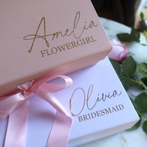 May include: Two gift boxes with pink and white lids. The top box is pink with gold lettering that says "Amelia Flowergirl". The bottom box is white with gold lettering that says "Olivia Bridesmaid". A pink ribbon is tied around the boxes.
