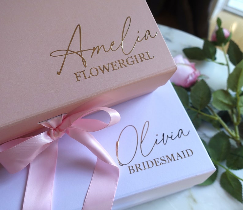 May include: Two gift boxes with pink and white lids. The top box is pink with gold lettering that says "Amelia Flowergirl". The bottom box is white with gold lettering that says "Olivia Bridesmaid". A pink ribbon is tied around the boxes.
