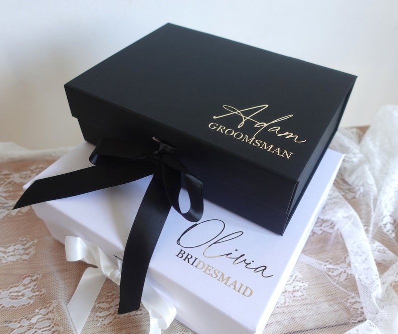 May include: Two gift boxes, one black and one white, with gold lettering. The black box says "Adam Groomman" and the white box says "Olivia Bridesmaid". Both boxes are tied with black ribbon.