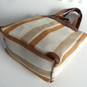 May include: A striped tote bag with brown leather handles. The bag is made of a woven fabric with white, beige, and orange stripes.