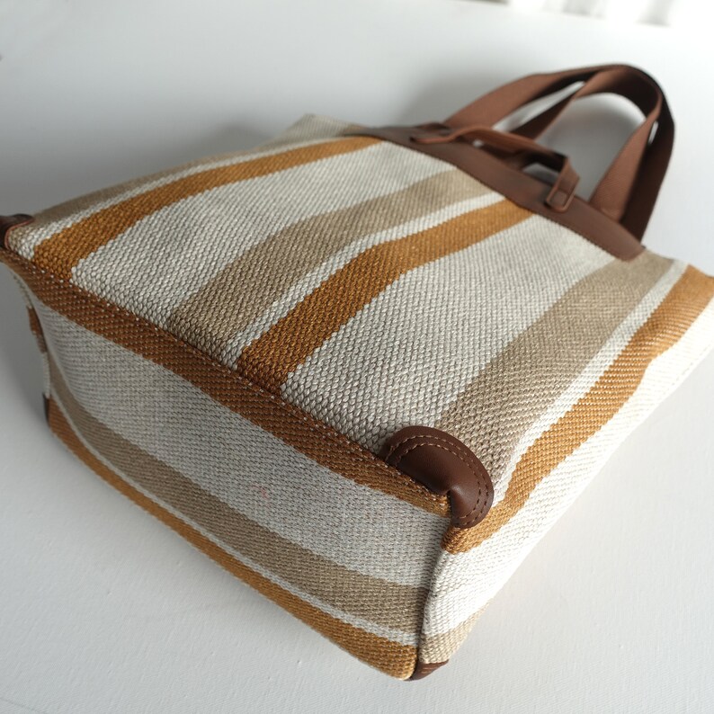 May include: A striped tote bag with brown leather handles. The bag is made of a woven fabric with white, beige, and orange stripes.