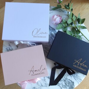 May include: Three gift boxes with personalised names and titles. The boxes are white, pink and black with matching ribbon. The names are Olivia Bridesmaid, Amelia Flower Girl, and Adam Groomsman.