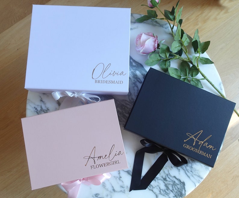 May include: Three gift boxes with personalised names and titles. The boxes are white, pink and black with matching ribbon. The names are Olivia Bridesmaid, Amelia Flower Girl, and Adam Groomsman.