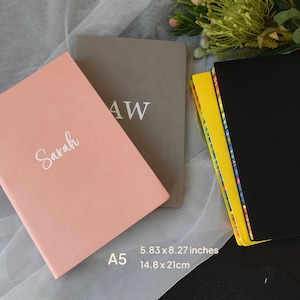 May include: Three notebooks with different covers. The first notebook is pink with the name "Sarah" written in white. The second notebook is grey with the letters "AW" written in white. The third notebook is black with a yellow and colourful striped border. The notebooks are all A5 size and measure 5.83 x 8.27 inches or 14.8 x 21 cm.