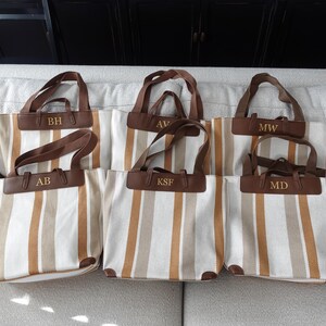 May include: Six striped canvas tote bags with brown leather handles and personalized initials. The initials are "BH", "AB", "AV", "KSF", "MW", and "MD".