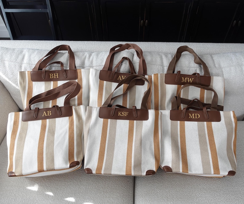 May include: Six striped canvas tote bags with brown leather handles and personalized initials. The initials are "BH", "AB", "AV", "KSF", "MW", and "MD".