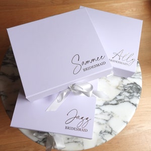 May include: Three white gift boxes with silver lettering. The boxes are tied with white satin ribbon. The boxes are labelled "Sammee Bridesmaid", "Ally Bridesmaid", and "Jazz Bridesmaid".