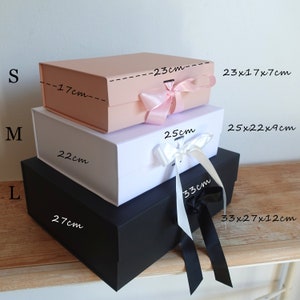 May include: Three gift boxes stacked on top of each other. The top box is pink, the middle box is white, and the bottom box is black. The boxes are labelled with the letters S, M, and L, and the dimensions of each box are listed in centimetres. The boxes are tied with ribbon.
