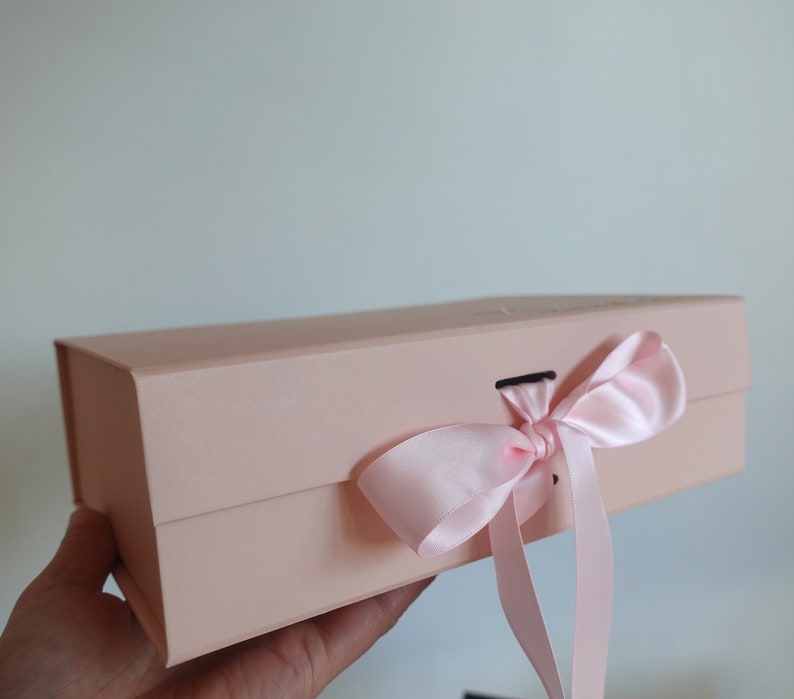 May include: A pale pink gift box with a satin ribbon tied in a bow. The box has a magnetic closure.