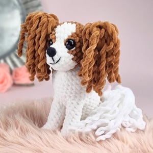 May include: A plush Cavalier King Charles Spaniel dog with brown and white fur. The dog has long, curly brown ears and a white patch on its head. It is sitting on a pink, fluffy surface.