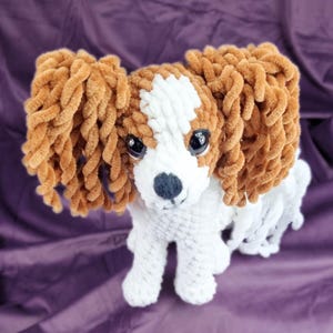 May include: A handmade plush Cavalier King Charles Spaniel dog. The toy dog has brown and white fur, with large, dark eyes and a black nose. The dog is made of soft, textured yarn and is standing on a purple surface.