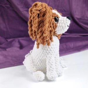 May include: A handmade, plush Cavalier King Charles Spaniel dog. The dog is white with brown and tan accents, including long, curly fur around the ears. It has large, black eyes and a black nose. The dog is sitting upright on a white surface.