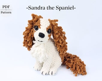 Könnte beinhalten: Ein gehäkeltes Cavalier King Charles Spaniel Hundespielzeug in Braun und Weiß. Der Hund sitzt mit seinem Schwanz hinter sich eingerollt. Das Spielzeug ist aus einem flauschigen Garn gefertigt und hat schwarze Augen und eine schwarze Nase.