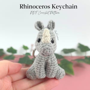 May include: A crocheted rhinoceros keychain in shades of gray and white. The keychain has a silver ring and is handmade. The text "Rhinoceros Keychain PDF Crochet Pattern" is at the top of the image.