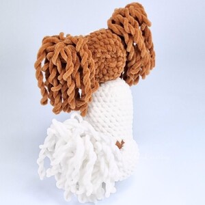 May include: A handmade, plush toy in the shape of a dog's head. The toy features a white body with a brown head and long, curly brown hair. The toy is made of soft, textured yarn.