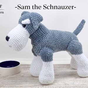 Könnte beinhalten: Ein gehäkeltes graues und weißes Miniatur-Schnauzer-Hundespielzeug. Der Hund steht auf einer weißen Oberfläche mit einer blauen und weißen Schüssel im Vordergrund. Der Text "Sam the Schnauzer" steht über dem Hund.