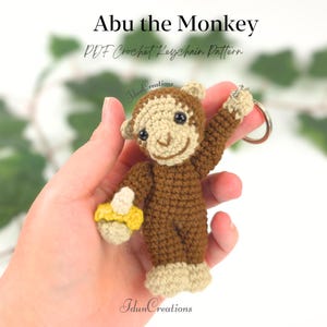 Pode incluir: Um chaveiro de macaco de crochê marrom e bege, segurando uma banana amarela. O chaveiro está preso a um anel prateado. O texto "Abu the Monkey PDF Crochet Keychain Pattern" é exibido na parte superior.