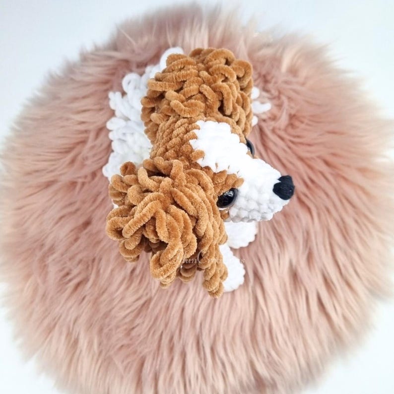 May include: A handmade, plush dog toy with brown, white, and black details. The dog's head is made of soft, textured yarn and is set against a pink, fluffy background. The toy has a realistic appearance, with a black nose and detailed eyes.