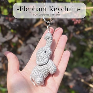 Op de afbeelding: Een gehaakte olifanten sleutelhanger in lichtgrijs, vastgehouden in een hand. De olifant heeft een slurf, oren en kleine zwarte ogen. De sleutelhanger is bevestigd aan een zilveren ring. Tekst op de afbeelding luidt "-Elephant Keychain- PDF Crochet Pattern."