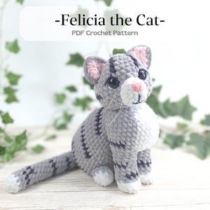 以下が含まれることがあります： グレーと白の毛並み、黒い目、ピンクの鼻と耳を持つ、フェリシアという名前のかぎ針編みの猫。猫は濃い灰色の縞模様があり、直立しています。「-Felicia the Cat- PDF Crochet Pattern」というテキストが上部にあります。