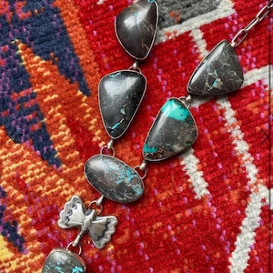 May include: A silver necklace featuring irregularly shaped, dark gray stones with turquoise accents. The necklace has a chain and a decorative butterfly pendant. The stones are connected by silver links, creating a unique and artistic design.